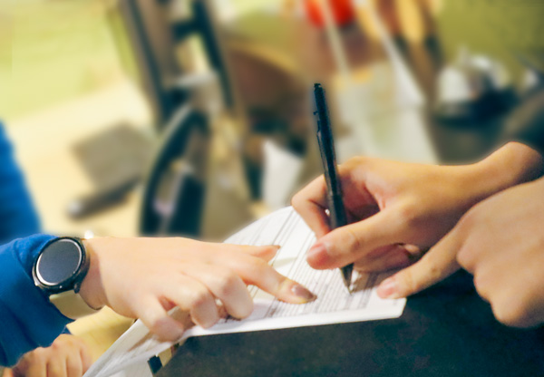 A hand points to a white paper while another hand holds a pen, signing it.