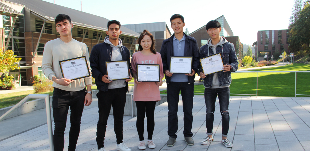 five students in a line holding scholarship award certificates with Cascadia College campus in the background